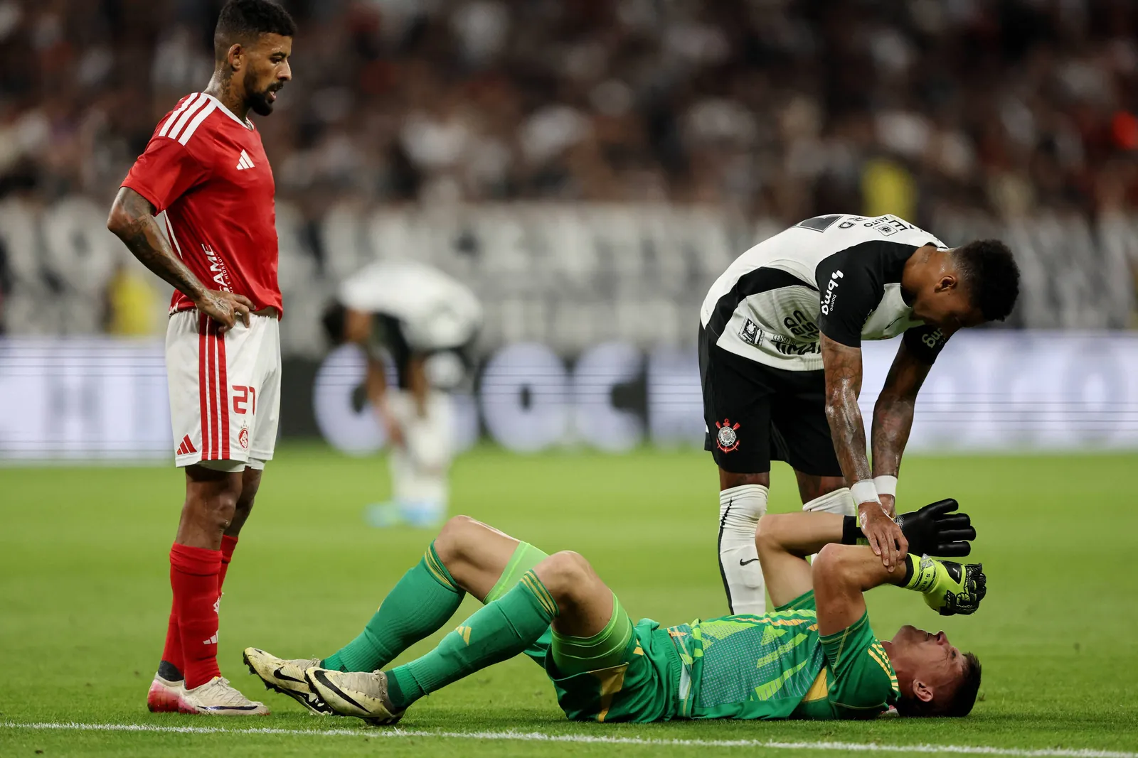Jogador de futebol vestido com uniforme verde estÃ¡ deitado no gramado segurando a cabeÃ§a, enquanto companheiro de time com uniforme preto e branco se inclina para ajudÃ¡-lo. Jogador adversÃ¡rio com uniforme vermelho observa a cena em pÃ©.