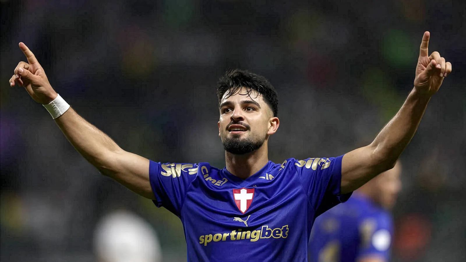 Jogador de futebol com uniforme azul do Cruzeiro levanta os braÃ§os em comemoraÃ§Ã£o, com expressÃ£o focada. Fundo desfocado com pÃºblico e campo.