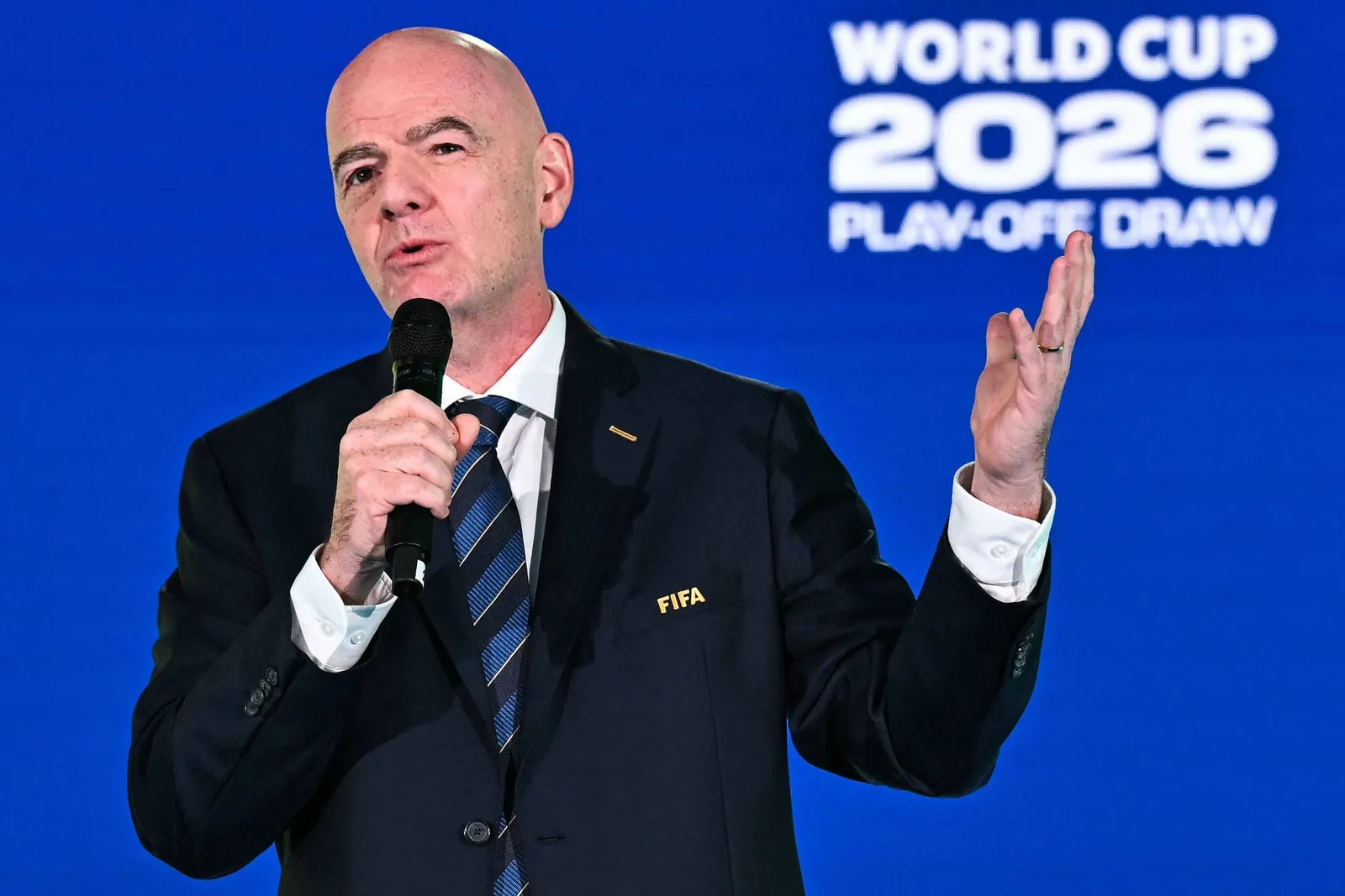 Homem de terno escuro com gravata listrada segura microfone e gesticula com a mão direita. Fundo azul exibe texto 'WORLD CUP 2026 PLAY-OFF DRAW'.