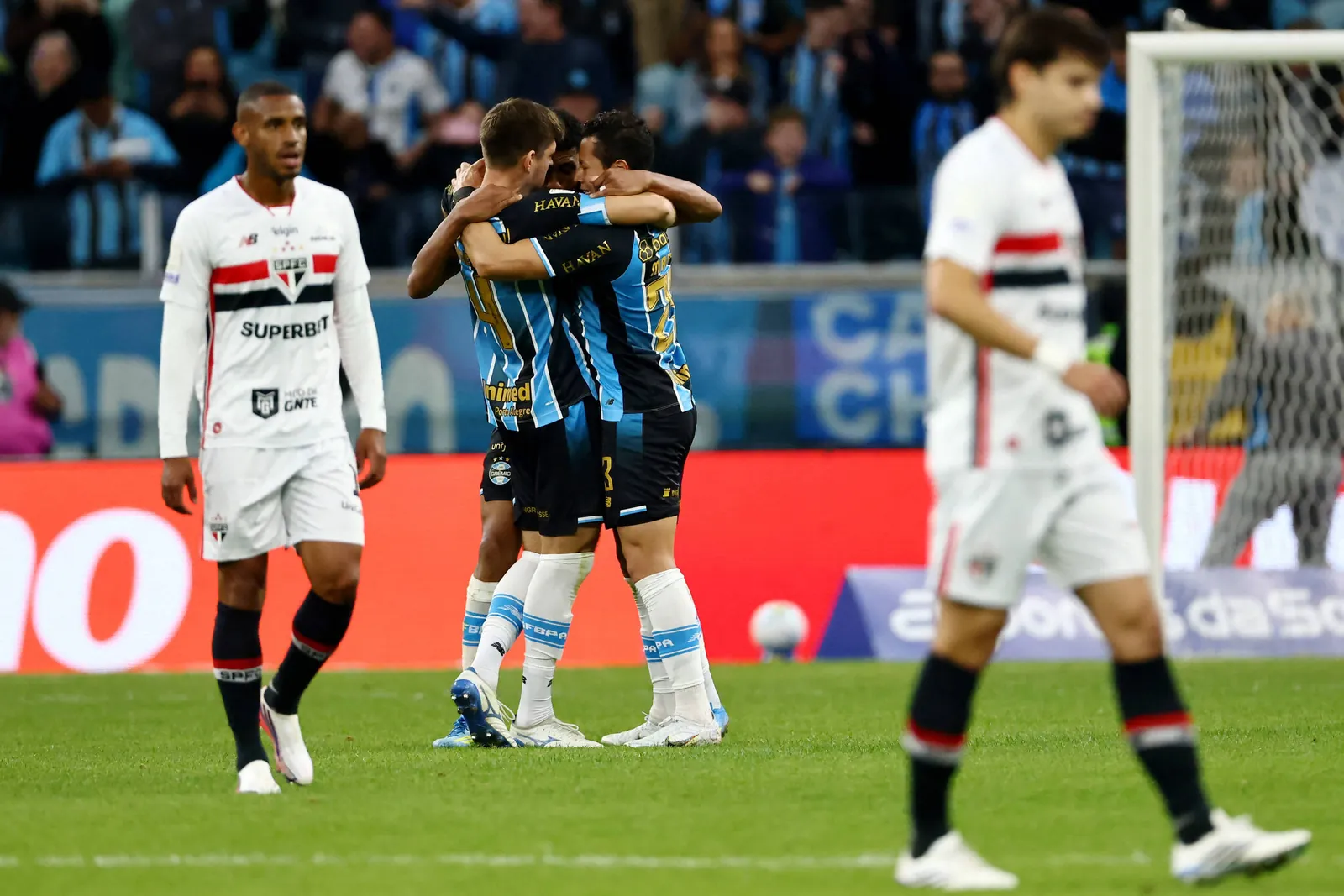 Dois jogadores do GrÃªmio abraÃ§am-se no centro do campo celebrando gol, enquanto jogadores do SÃ£o Paulo caminham cabisbaixos prÃ³ximos Ã Ã¡rea de gol. Torcida e placas publicitÃ¡rias aparecem ao fundo.